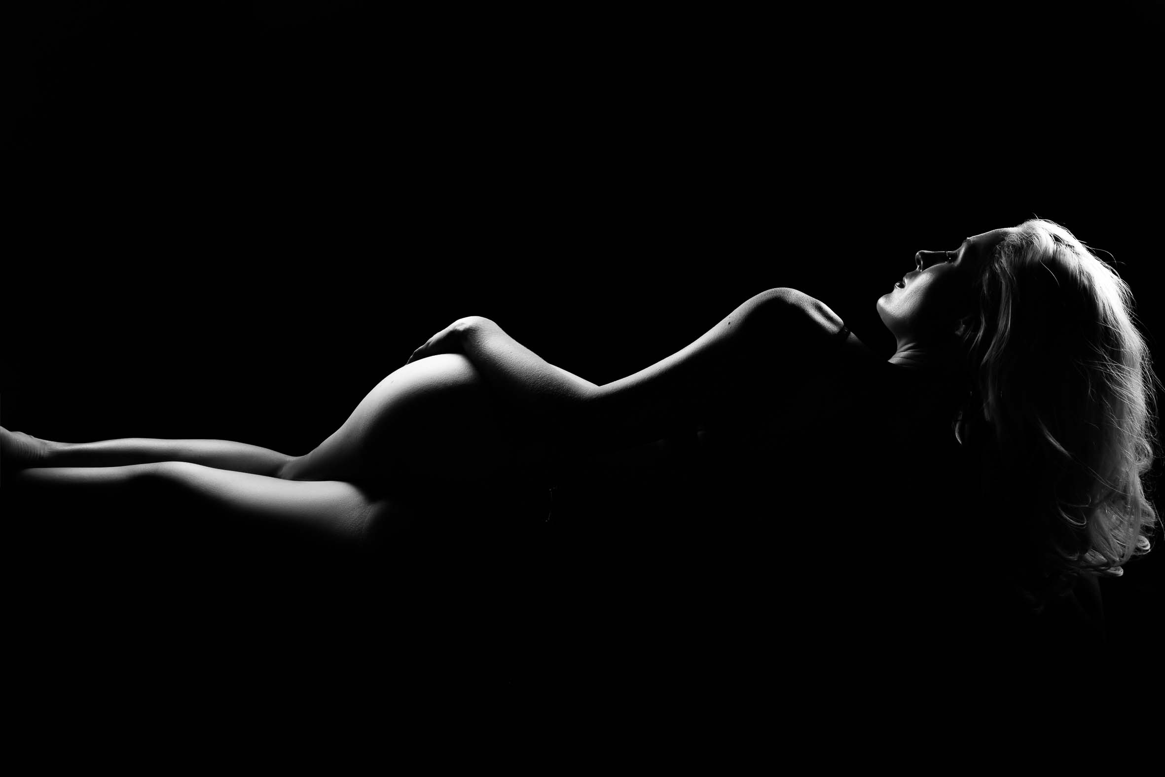 Black and white silhouette of a nude woman lying on her back with one arm resting on her hip and her head turned to the side, illuminated with dramatic lighting against a dark background.