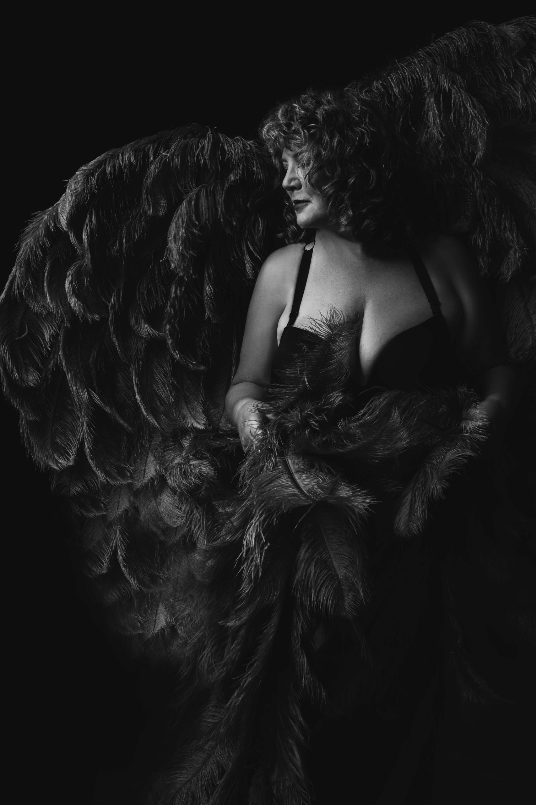 Black and white photo of a woman with curly hair wearing a black bra and draped in large dark feathers, looking to her left in a dark background.
