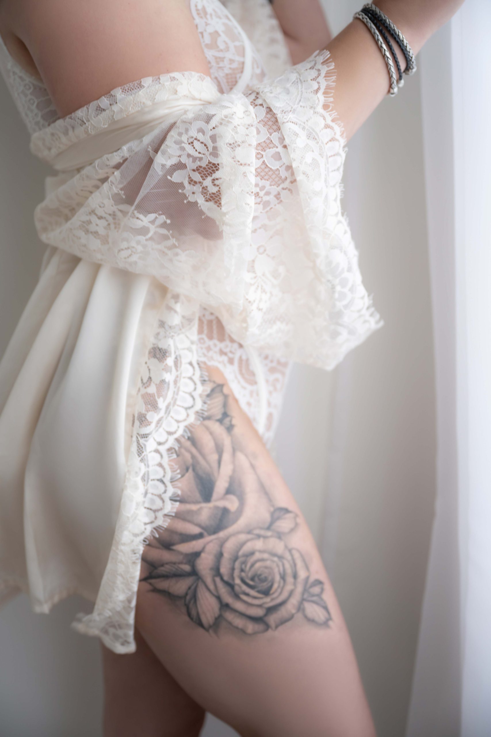 Close-up of a person's upper thigh with a detailed black and gray rose tattoo partially visible under white lace lingerie, wearing braided bracelets, standing near a white curtain.