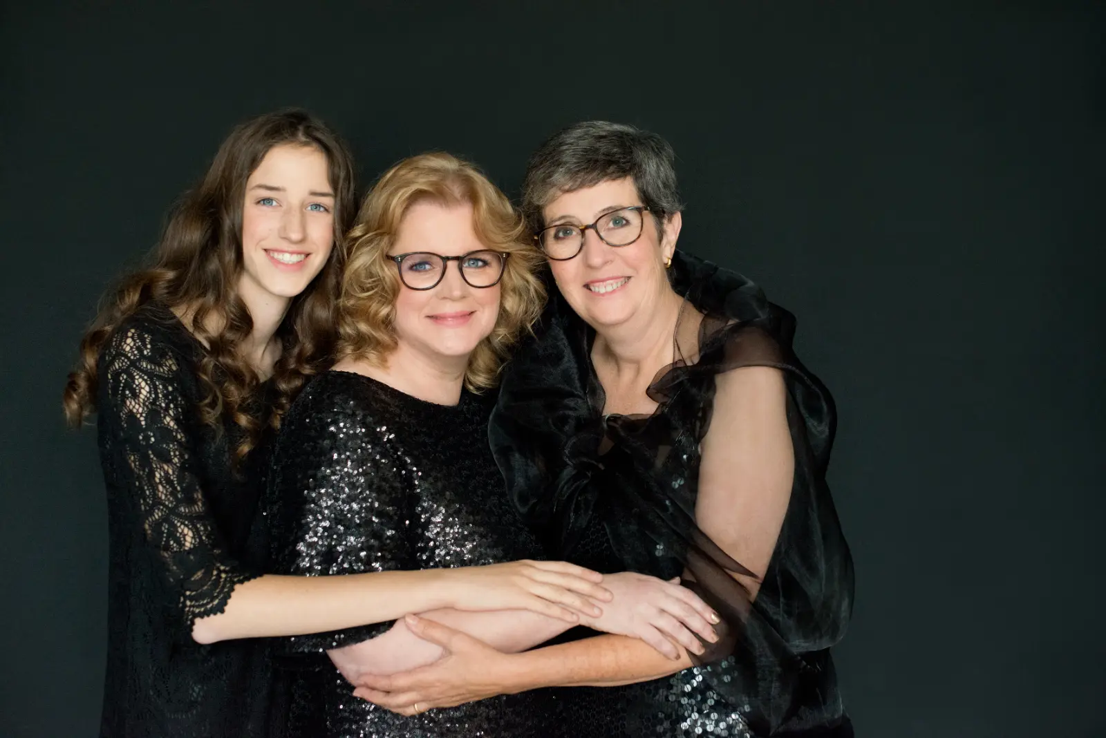 Three women of different ages in black dresses posing against a dark background, smiling and embracing each other warmly.
