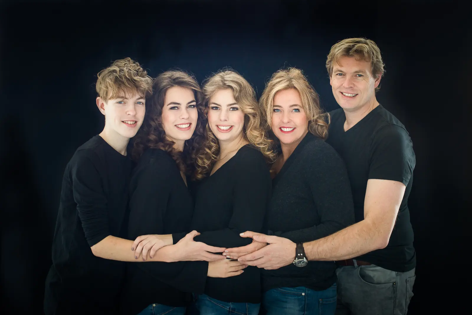 Five people standing closely in a group against a black background, smiling and embracing each other, wearing mostly black tops and blue jeans.