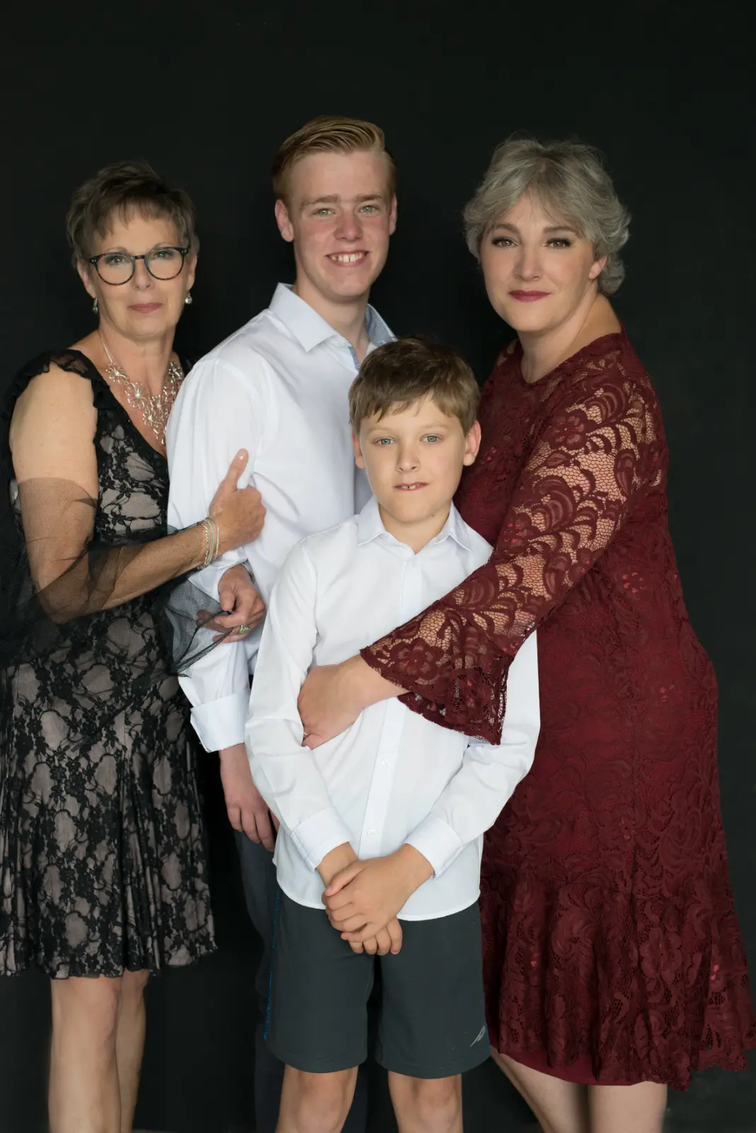 Portrait of four people posed against black background: a woman in glasses and black lace dress, a young man in a white shirt smiling, a woman in a burgundy lace dress hugging a young boy in a white shirt and dark shorts in front of her.