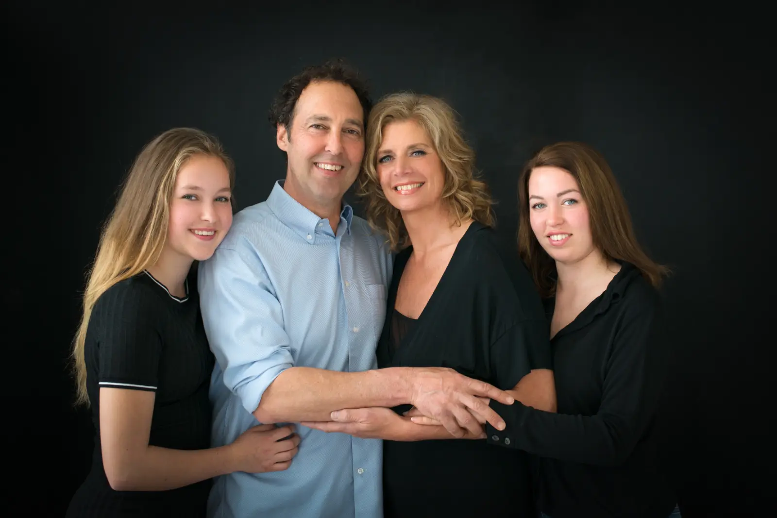 Portrait of a smiling family of four standing close together in front of a black background. A man in a light blue button-up shirt stands between two women in black tops, with a young woman in a black short-sleeve shirt on the left. They are embracing each other warmly.