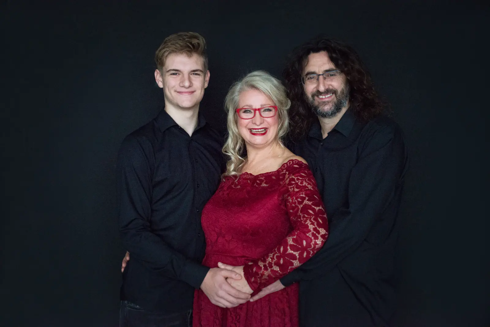 Smiling woman in red lace dress and red glasses standing between and embraced by two smiling men in black shirts against a black background.