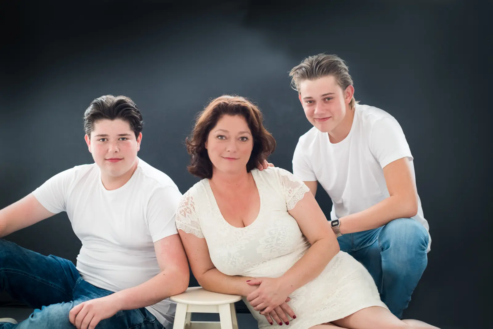 Portrait of a woman in a white lace dress leaning on a stool, flanked by two teenage boys wearing white t-shirts and jeans, against a dark background.