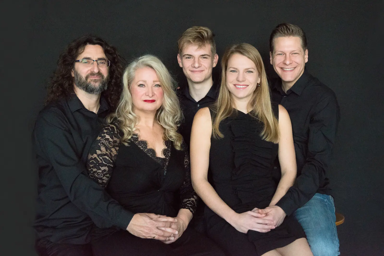 A group portrait of five people against a dark background: three men and two women sitting closely, all wearing black clothing and smiling at the camera.