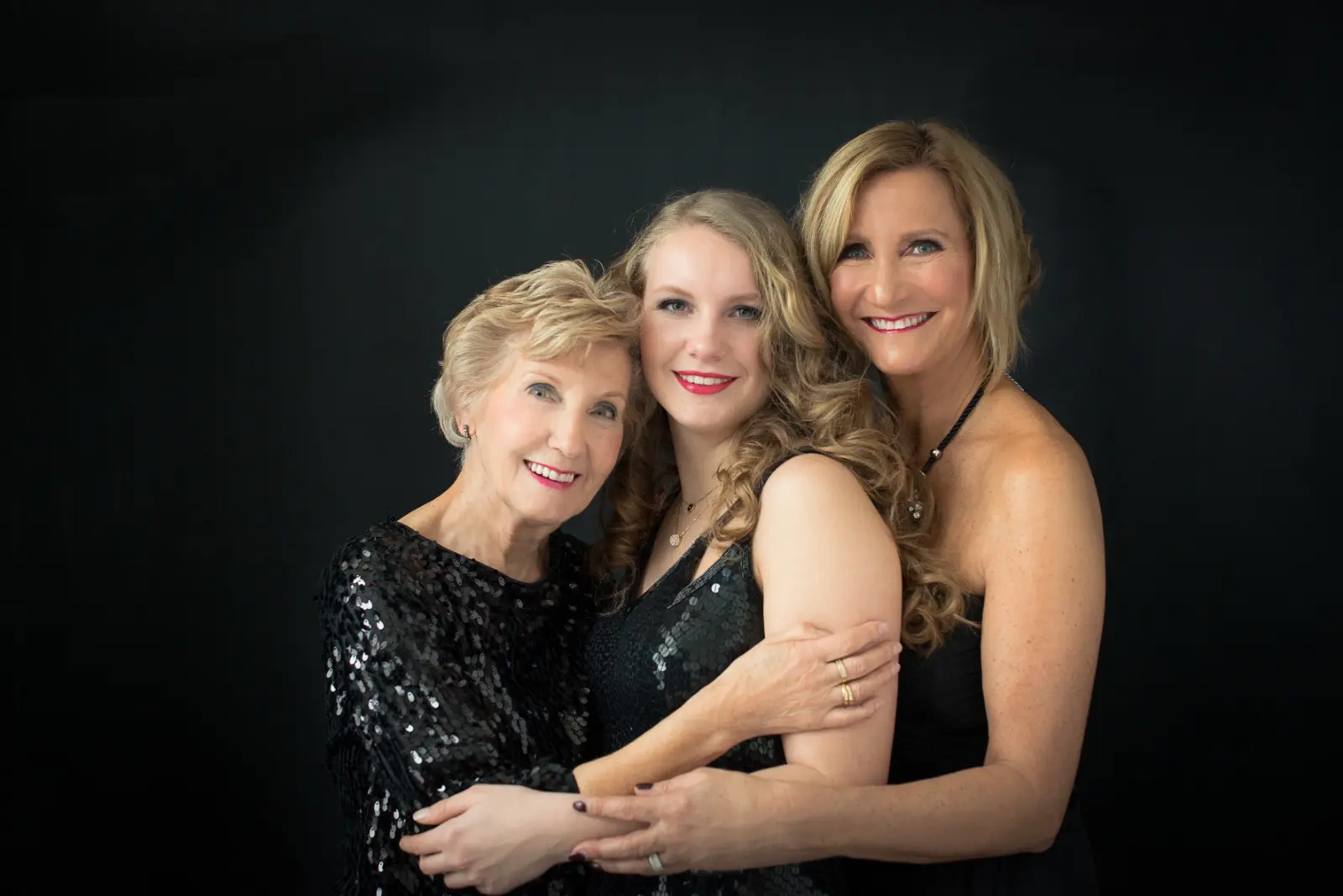 Three women of different ages stand closely together against a black background, smiling. The older woman on the left has short blonde hair and wears a black sequined top. The young woman in the center with long curly blonde hair wears a sleeveless black dress with sequins. The woman on the right has shoulder-length blonde hair and wears a sleeveless black dress, with their arms around each other.
