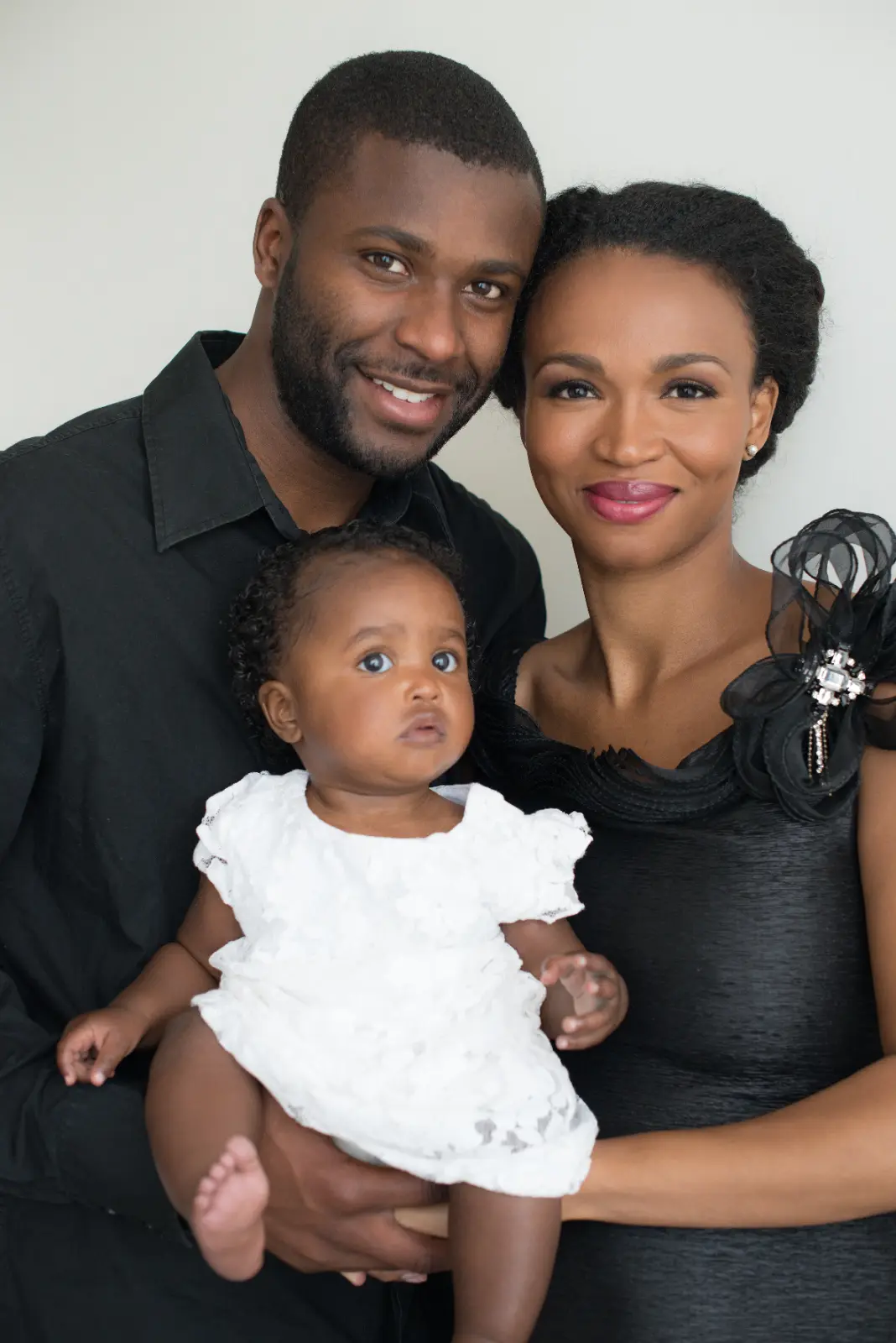 A smiling man and woman hold a baby dressed in a white outfit, posing together against a plain light background.