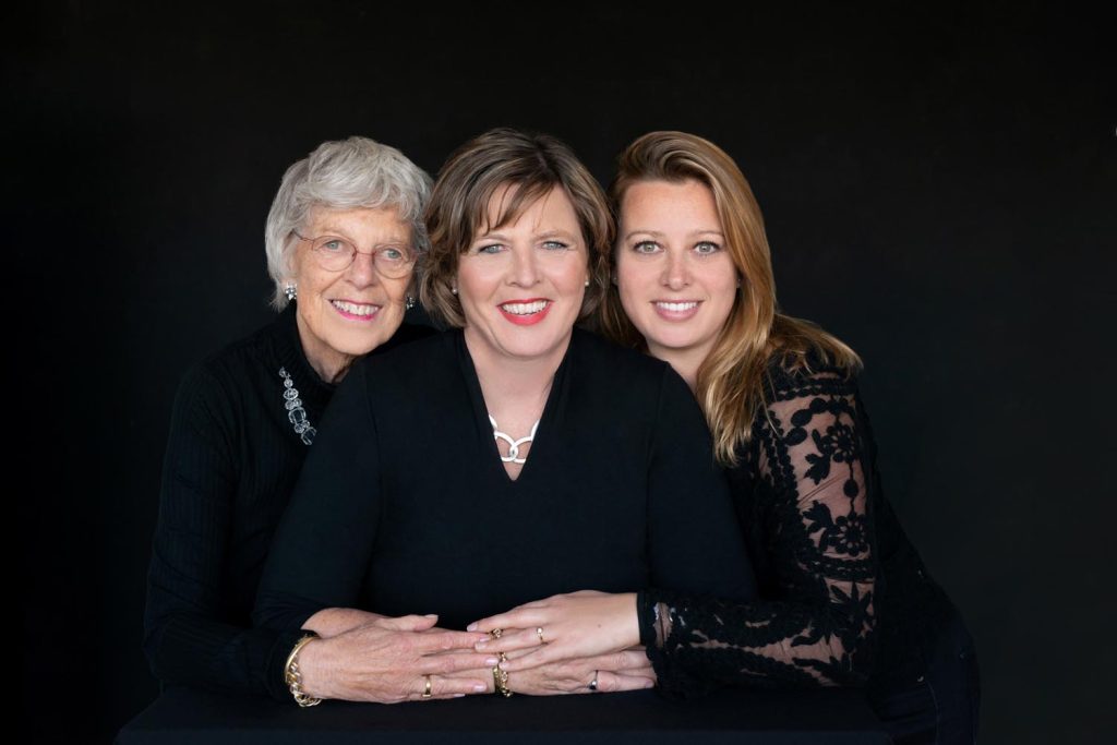 Three women of different generations smiling in a portrait against a black background, with the older woman on the left, a middle-aged woman in the center, and a younger woman on the right, all wearing black tops and closely embracing each other.