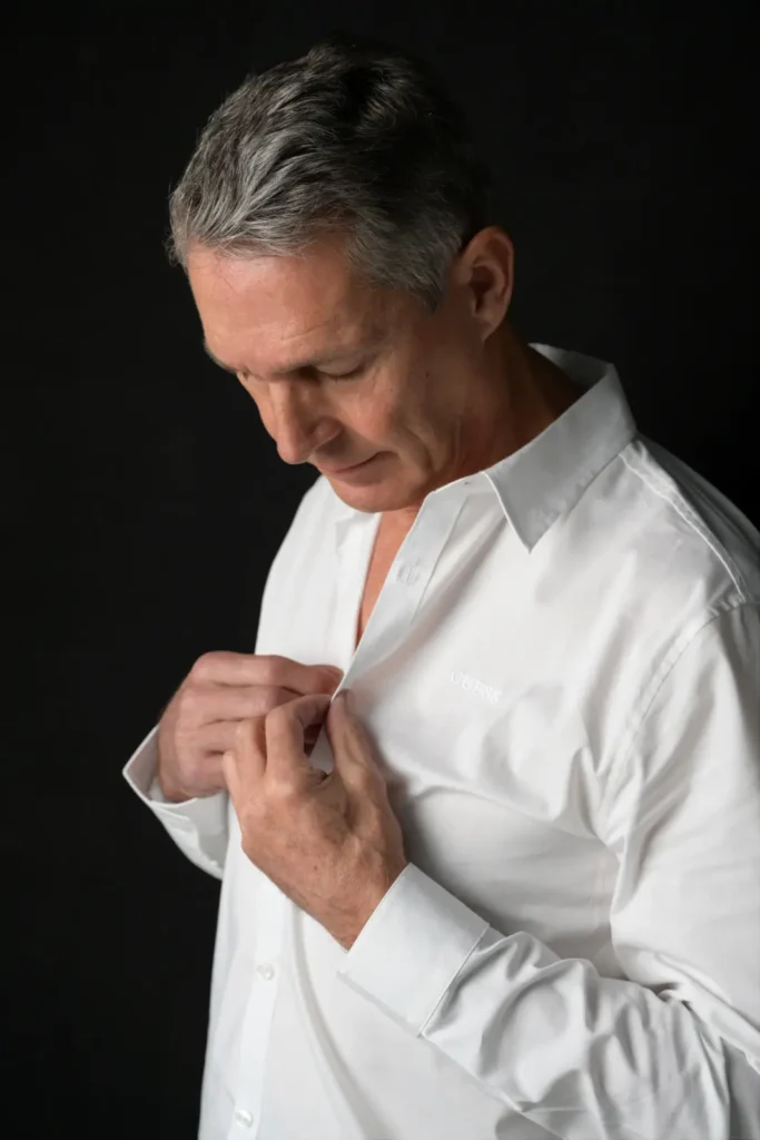 Middle-aged man with gray hair looking down while buttoning a white dress shirt against a black background.