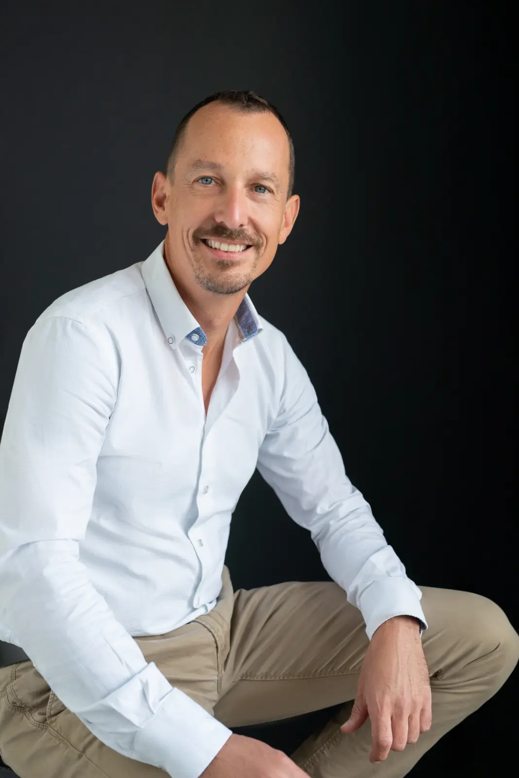 Smiling man with short hair and a trimmed beard wearing a white button-up shirt and beige pants sitting against a black background.