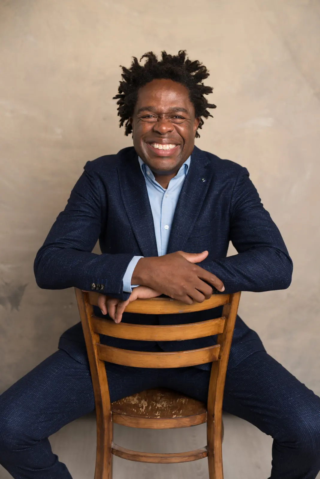 Smiling man with natural curly hair wearing a navy blue suit and light blue shirt sitting backward on a wooden chair against a plain beige background.