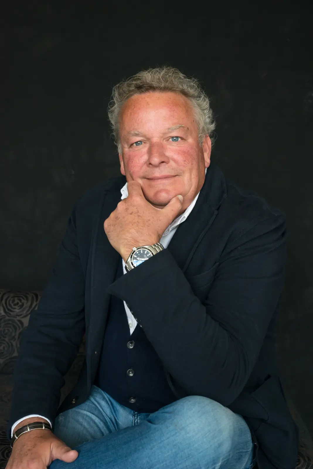 Middle-aged man with curly gray hair and blue eyes, wearing a dark blazer over a white shirt and dark vest, sitting with one hand resting on his chin and the other on his knee, against a dark background.