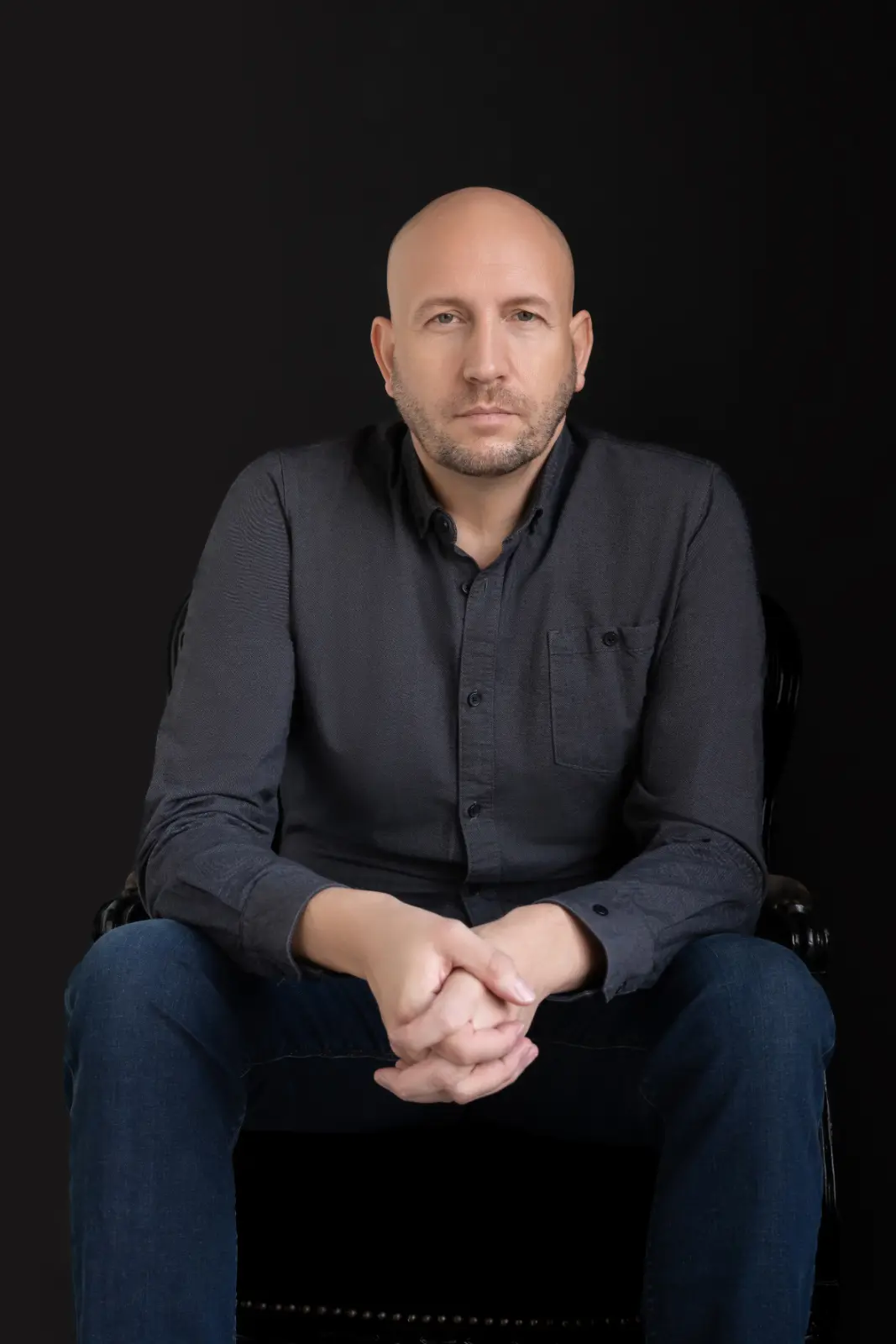 Portrait of a serious bald man with a beard wearing a dark shirt and blue jeans, sitting with hands clasped in front of a black background.