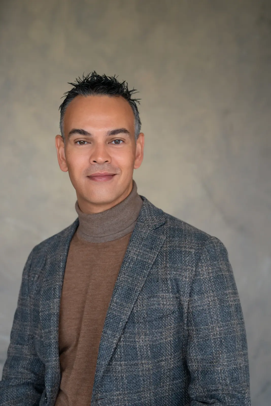 Portrait of a man with short, dark, spiked hair wearing a gray plaid blazer and a brown turtleneck sweater against a neutral background.