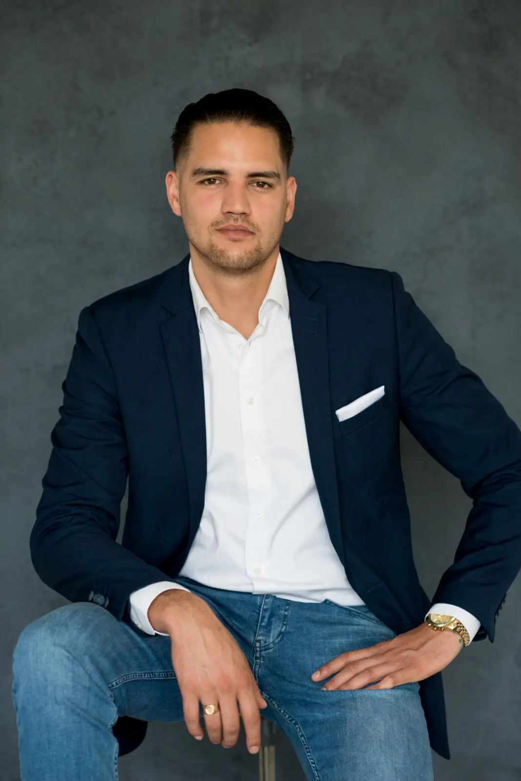 Confident man with slicked-back hair wearing a navy blazer, white dress shirt, light blue jeans, gold watch, and gold ring sitting with one hand on his knee and the other on his hip against a dark gray background.