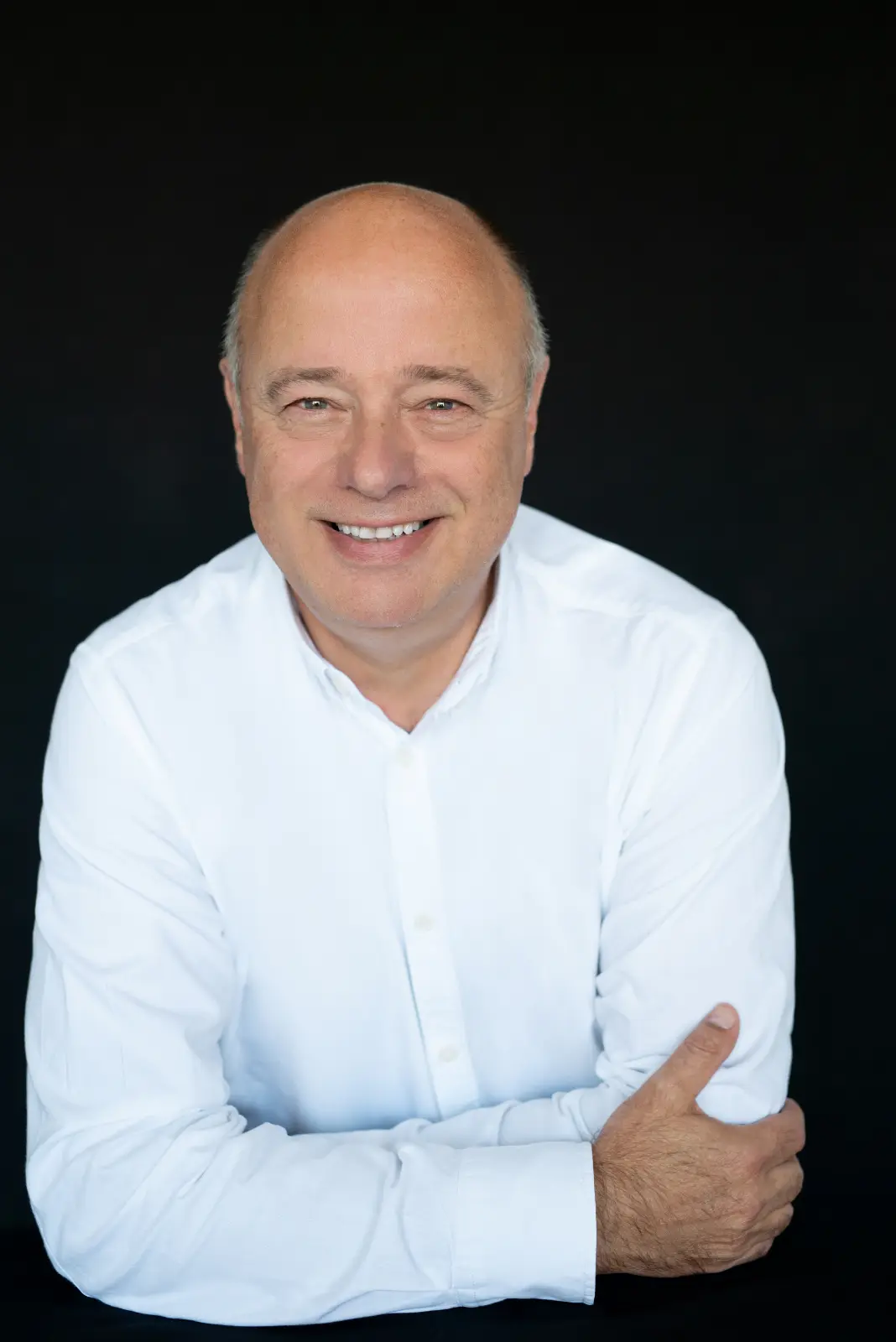 Smiling middle-aged bald man with light skin wearing a white button-up shirt, posed with arms crossed against a black background.