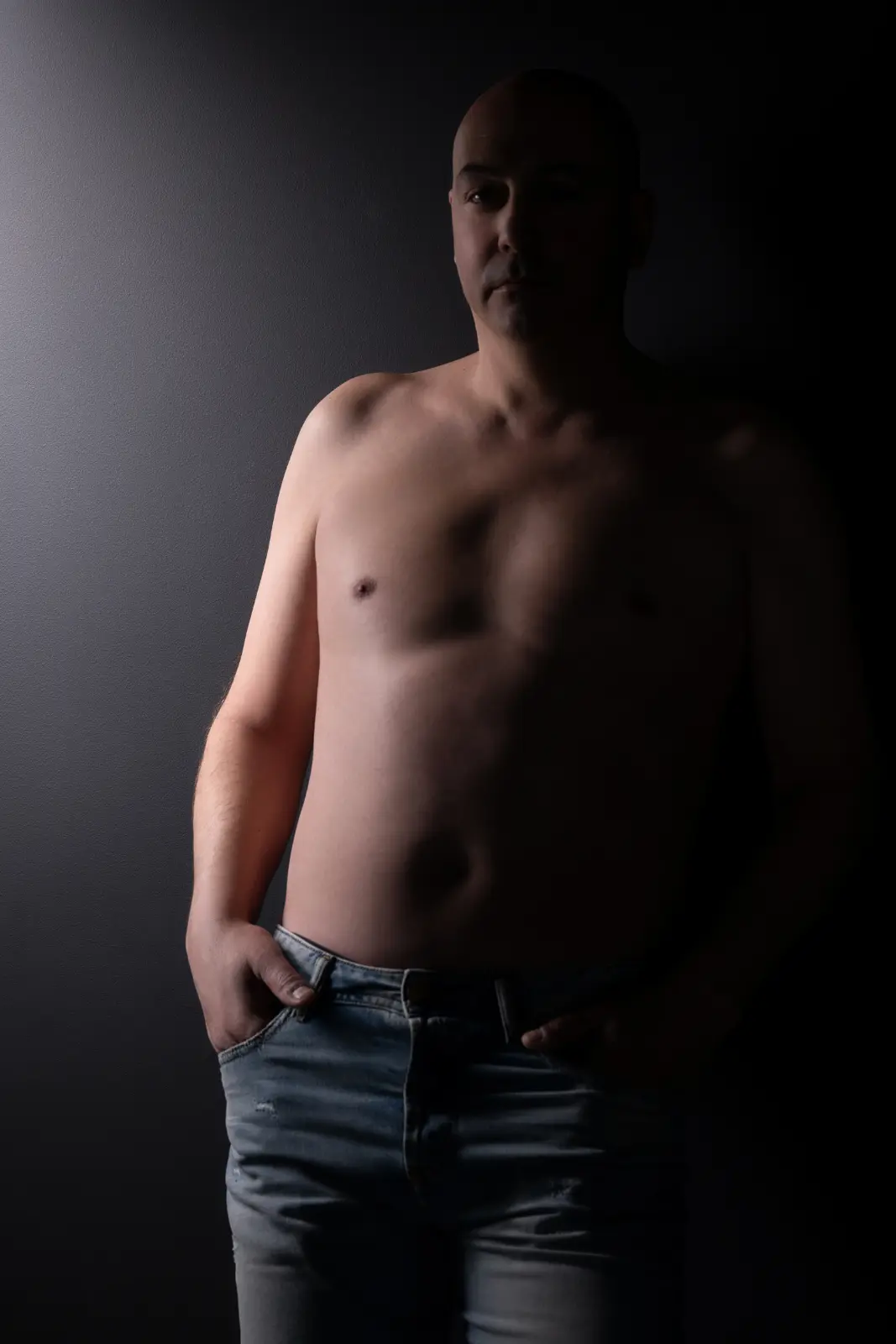 Shirtless man standing against a dark background with his hands in the pockets of his light blue jeans, illuminated from the left side creating shadows on his face and body.