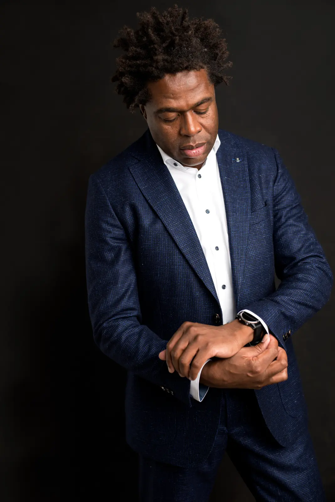 Man wearing a dark blue suit and white shirt looking down while adjusting his cuff above a smart watch on his wrist against a black background.