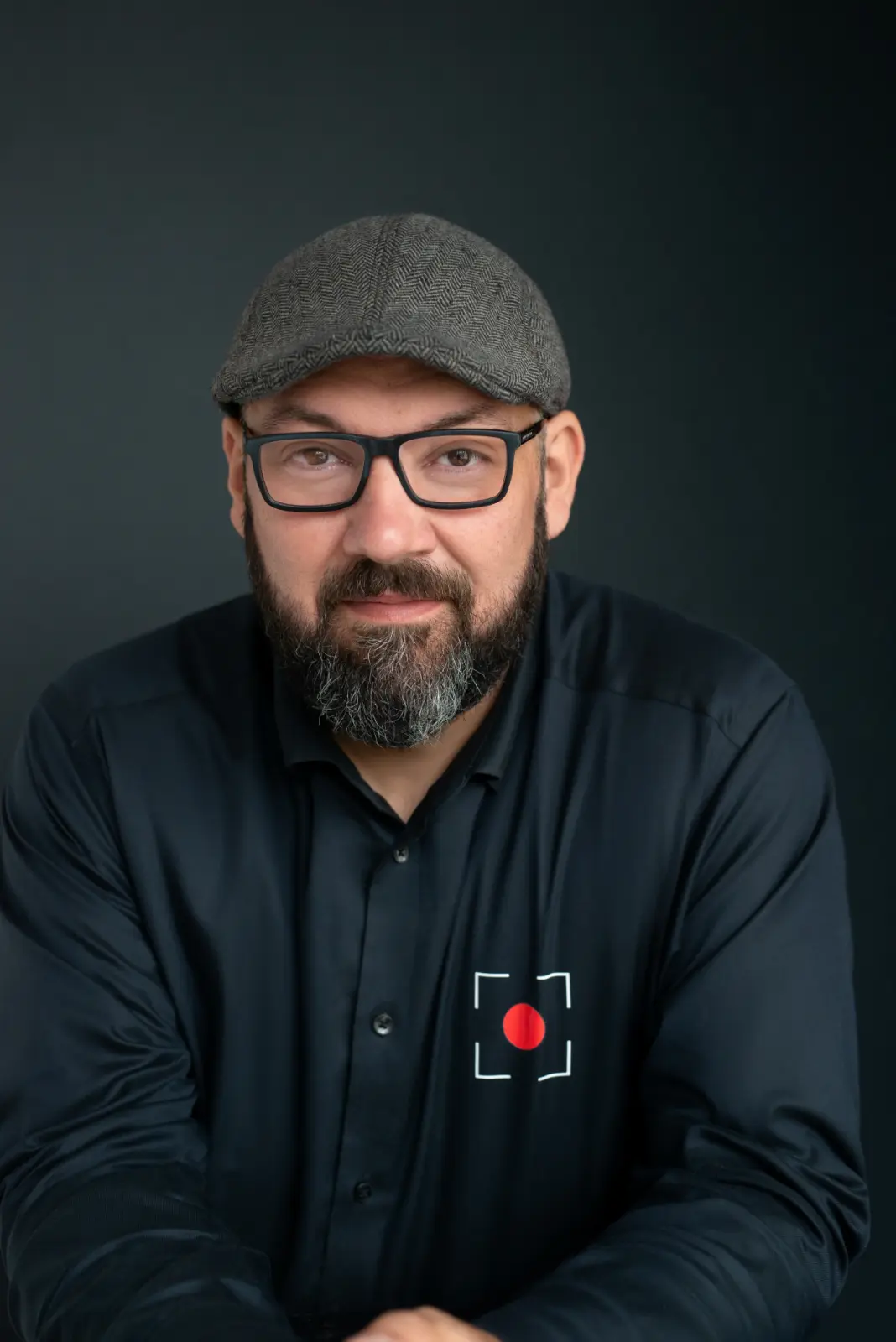 Middle-aged man with a full beard and glasses wearing a gray flat cap and black button-up shirt with a red dot and square frame design on the chest, posing against a dark background.