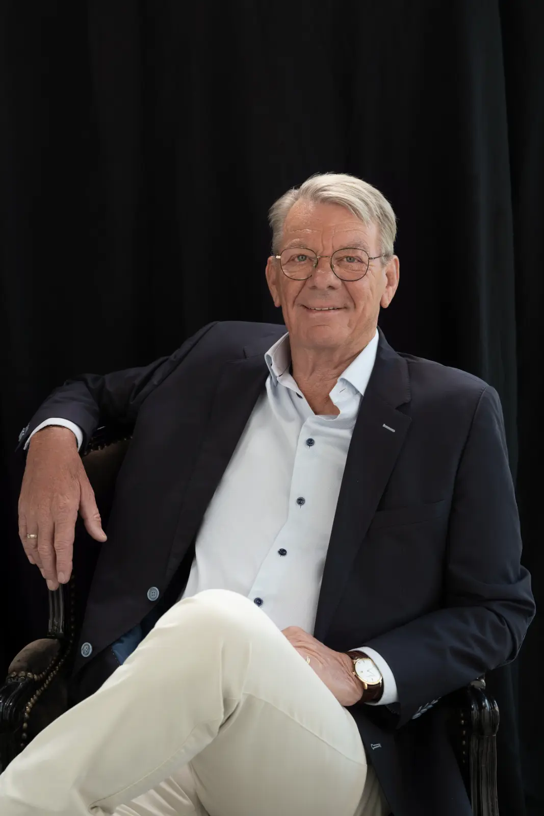 Elderly man with gray hair and glasses wearing a dark blazer, light blue shirt, beige pants, and a wristwatch, sitting on a black armchair against a black backdrop, smiling at the camera.