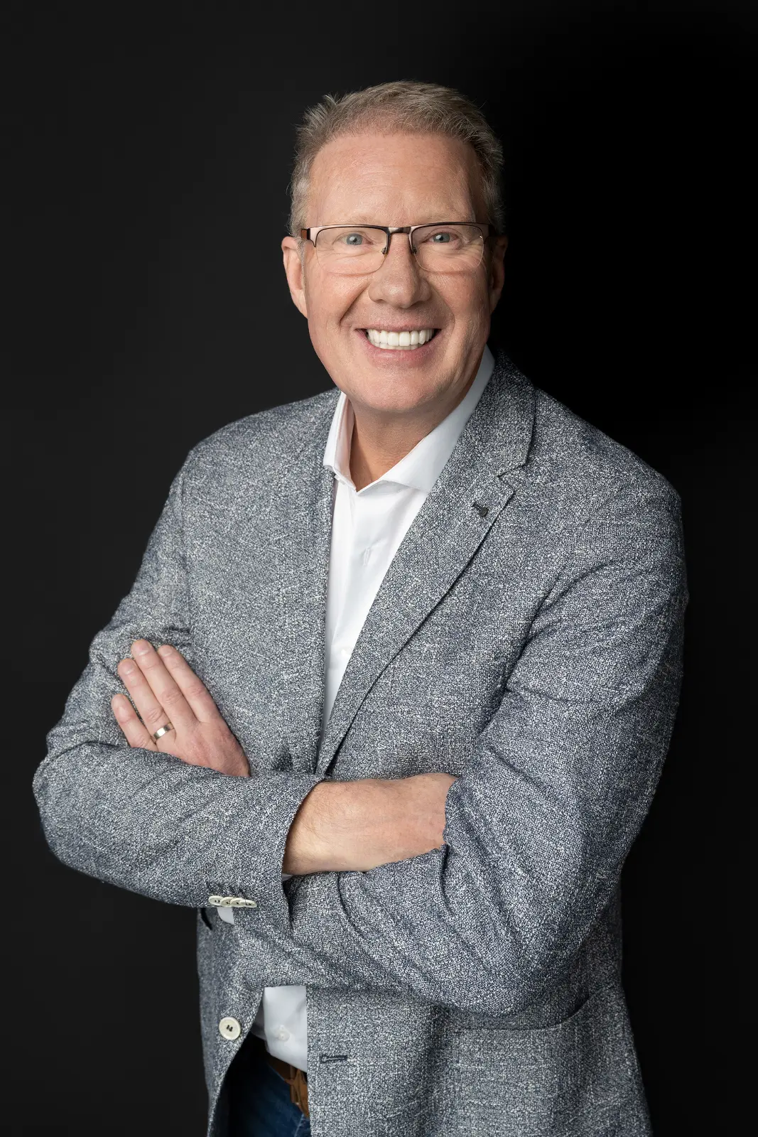 Smiling middle-aged man wearing glasses, a gray textured blazer, and a white shirt, standing with arms crossed against a black background.