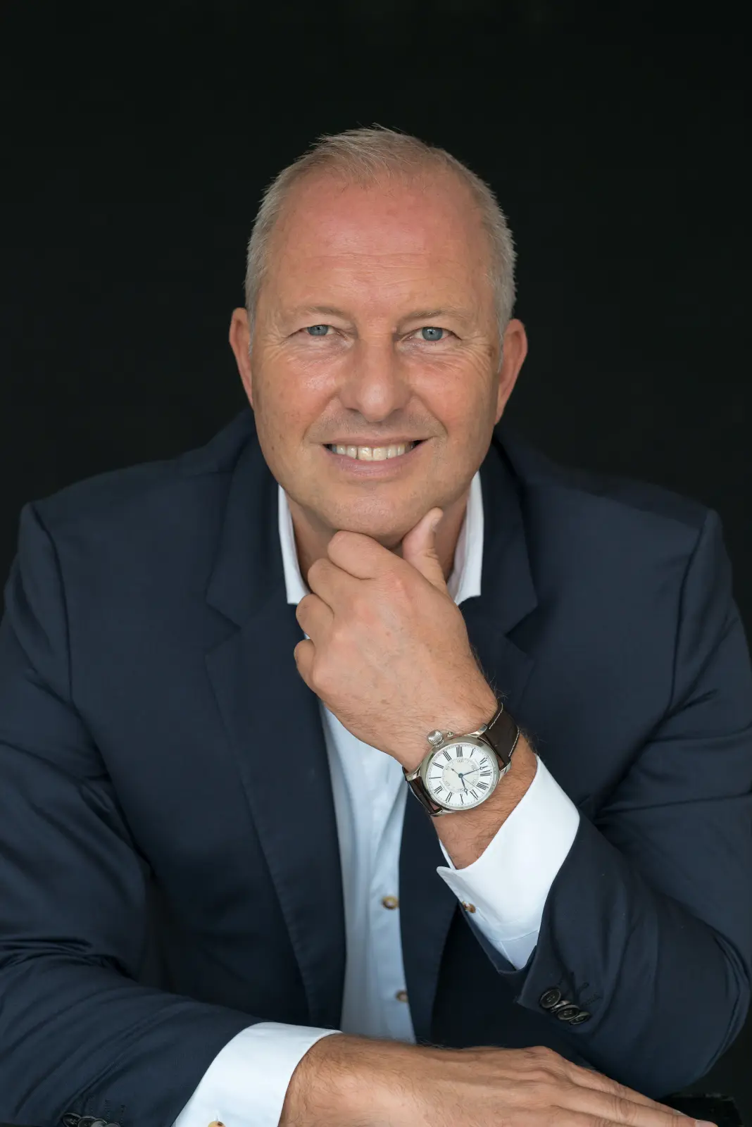 Smiling middle-aged man in a dark blazer and white shirt with a wristwatch, resting his chin on his hand against a black background.