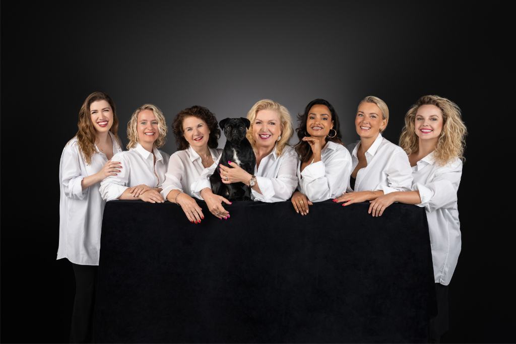 Seven women dressed in white shirts pose smiling behind a black cloth, with a small black dog held by the woman in the center against a dark background.