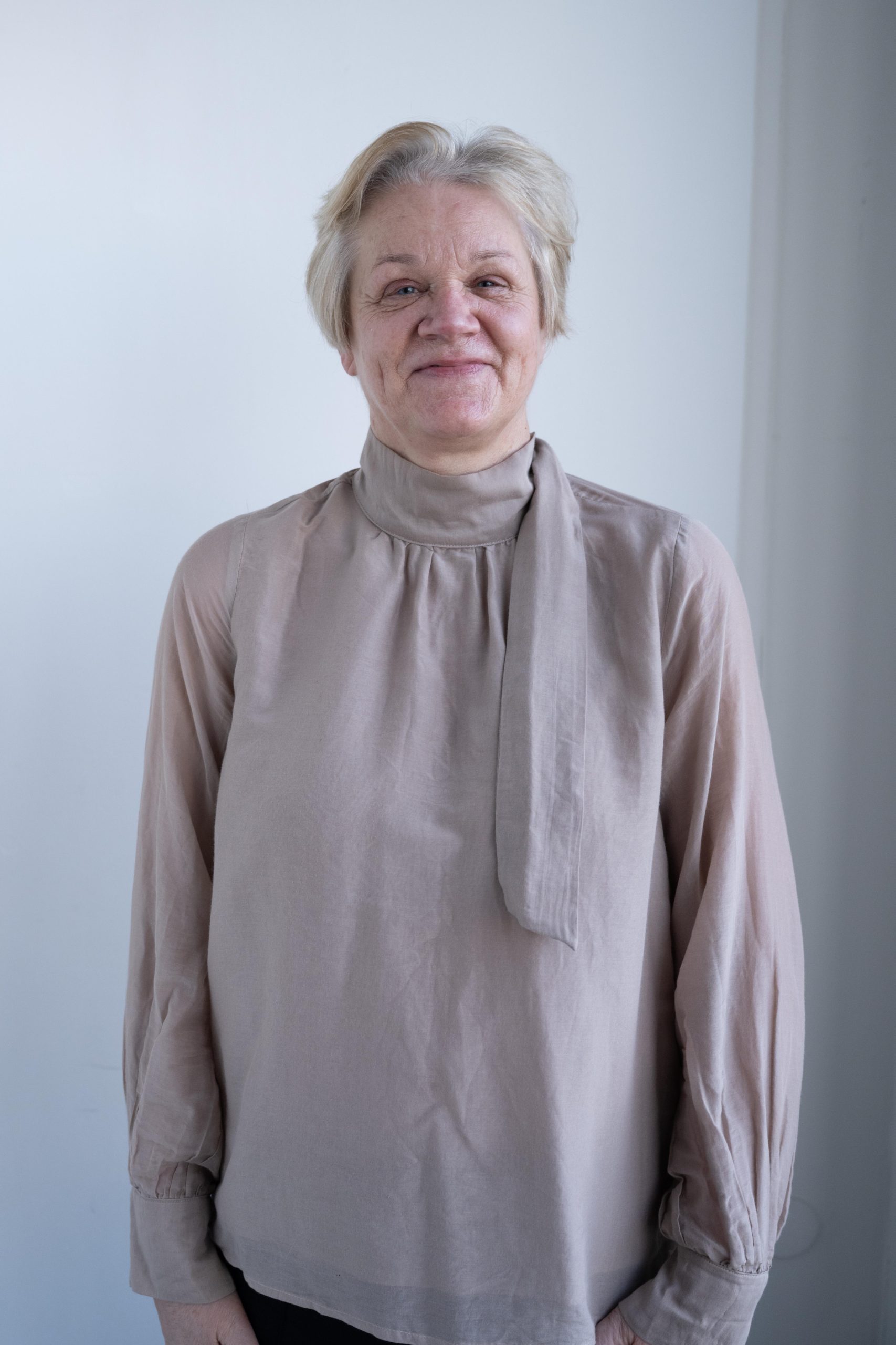 Portrait of a smiling older woman with short light blonde hair wearing a beige long-sleeve blouse with a loose scarf detail around the neck, standing against a plain light background.