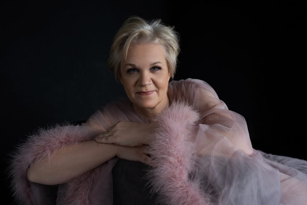 Portrait of a mature woman with short blonde hair, resting her chin on crossed arms and smiling gently, wearing a soft pink garment with fluffy trim against a dark background.