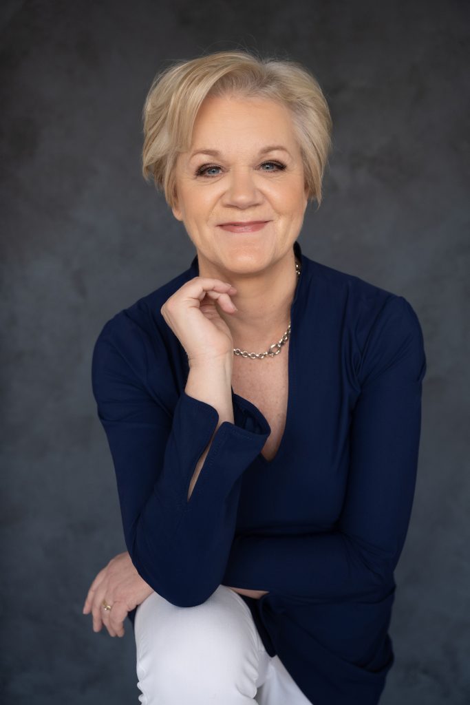 Smiling middle-aged woman with short blonde hair wearing a navy blue top and white pants, posing with her hand near her chin against a dark gray background.