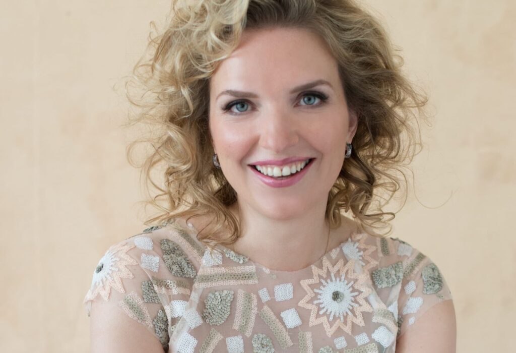 Smiling woman with curly blonde hair wearing a light, patterned top with sequins against a neutral background.