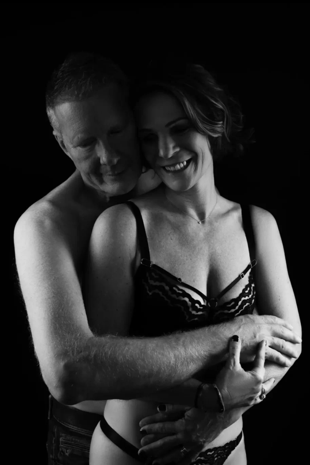 Black and white photo of a shirtless man embracing a smiling woman wearing black lace lingerie against a black background.