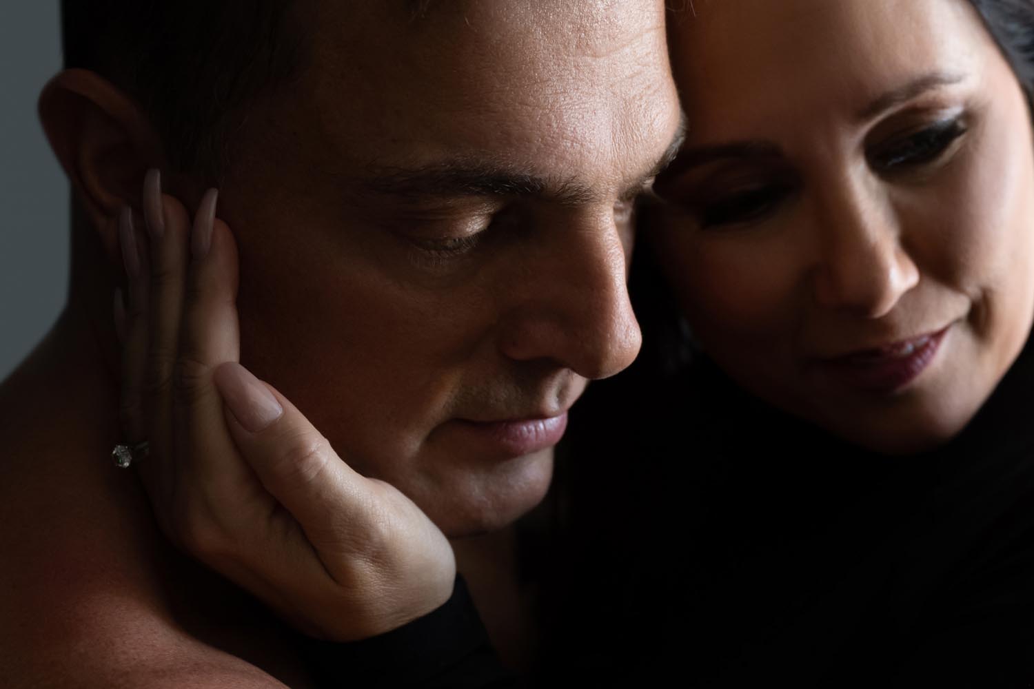 Close-up of a man and woman with their faces touching gently, the woman’s hand with an engagement ring resting on the man’s cheek in a dimly lit, intimate moment.