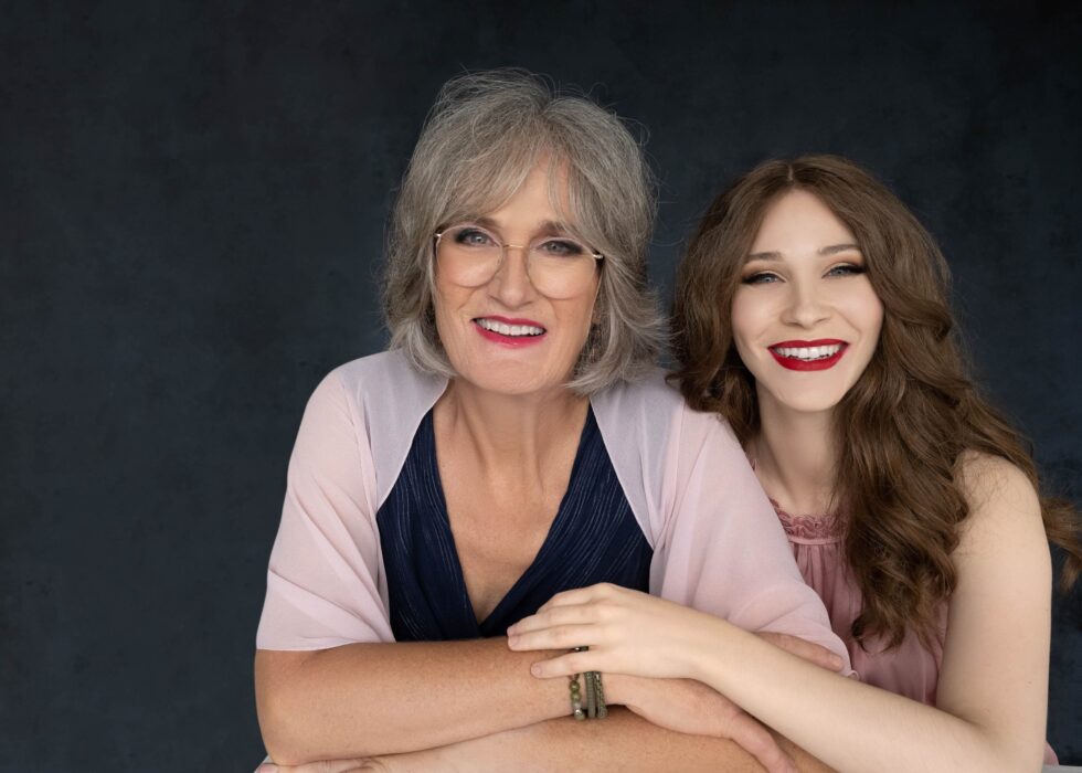 Smiling older woman with gray hair and glasses wearing a navy dress and light pink shawl, embraced by a younger woman with long curly brown hair and red lipstick, both posing against a dark gray background.