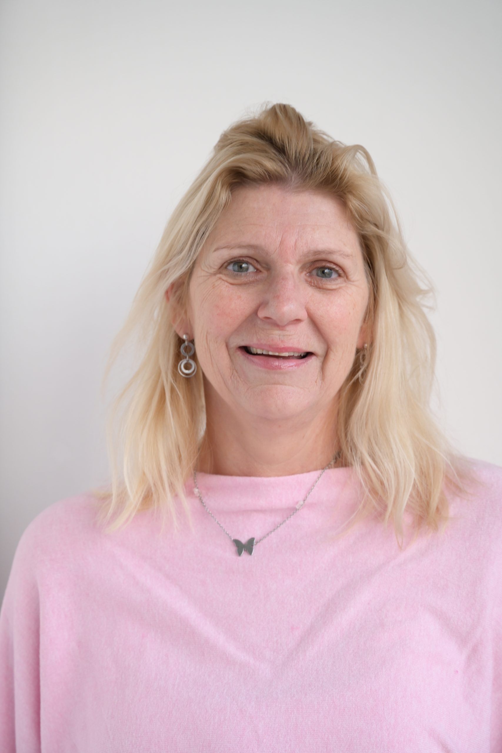 Blonde woman with light skin and blue eyes smiling, wearing silver hoop earrings, a butterfly necklace, and a light pink top, against a plain white background.