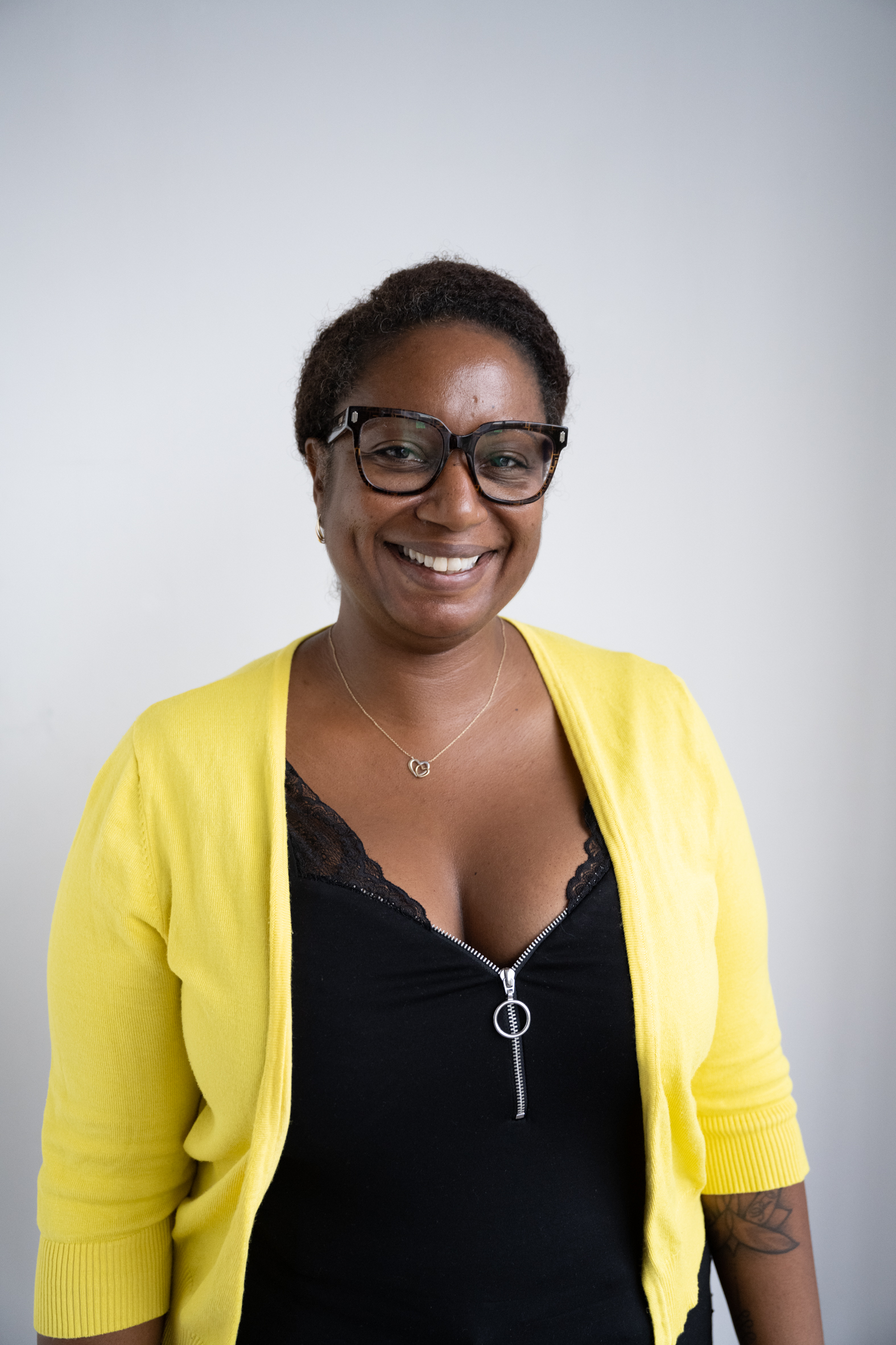 Smiling woman with short curly hair wearing large tortoiseshell glasses, a yellow cardigan over a black zippered top with lace trim, and a small double-heart necklace.
