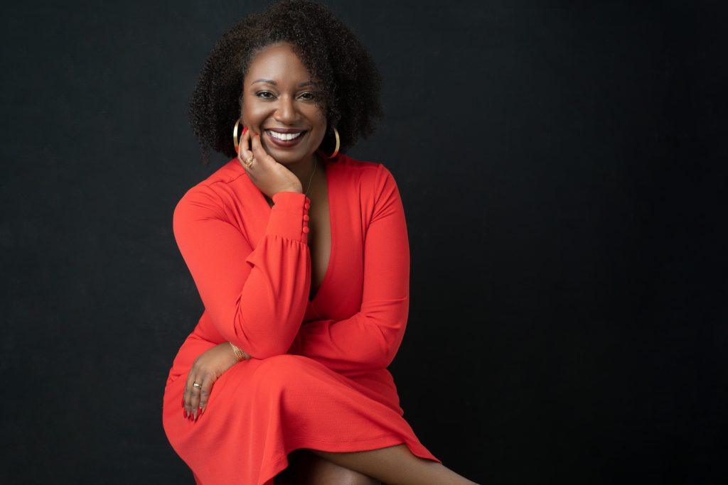 Smiling woman with curly hair dressed in a red long-sleeve dress sitting with one hand resting on her cheek and the other on her knee against a black background.