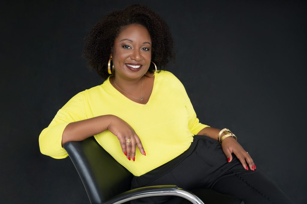 Smiling woman with curly hair wearing a bright yellow top and black pants, seated casually on a chair with a black background.