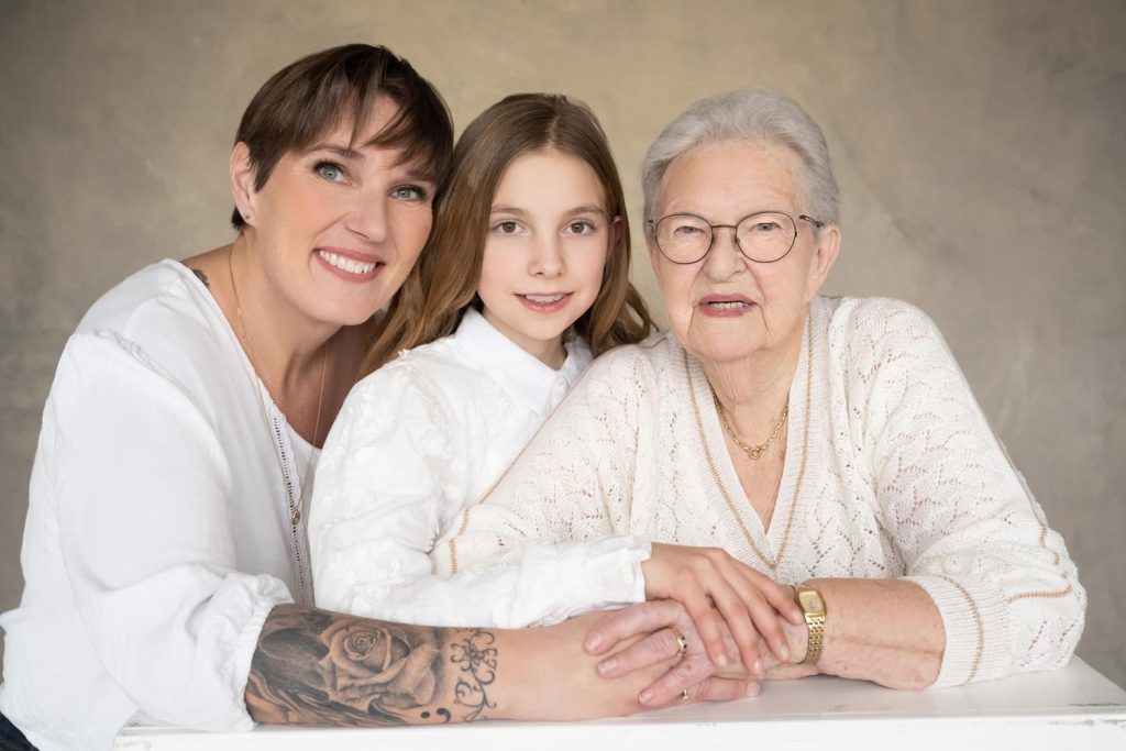 Portrait of three generations of women: an older woman with glasses and gray hair, a middle-aged woman with short hair and a tattoo on her arm, and a young girl with long brown hair, all wearing white tops and smiling gently while leaning on a white surface.