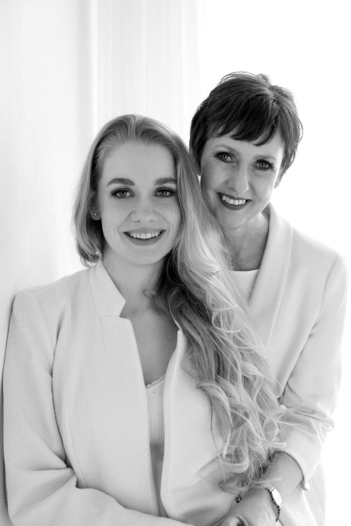 Black and white portrait of a smiling middle-aged woman embracing a younger woman with long curly hair, both wearing light-colored clothing, standing in front of a bright background.