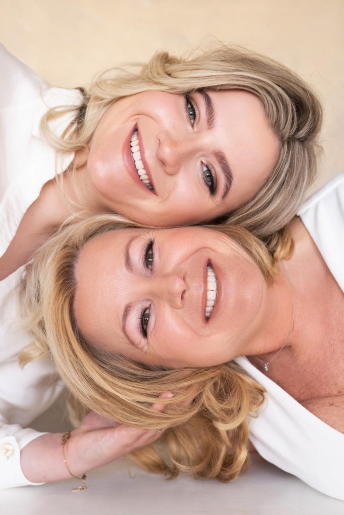 Two smiling blonde women lying close together with their heads touching, both wearing white tops.