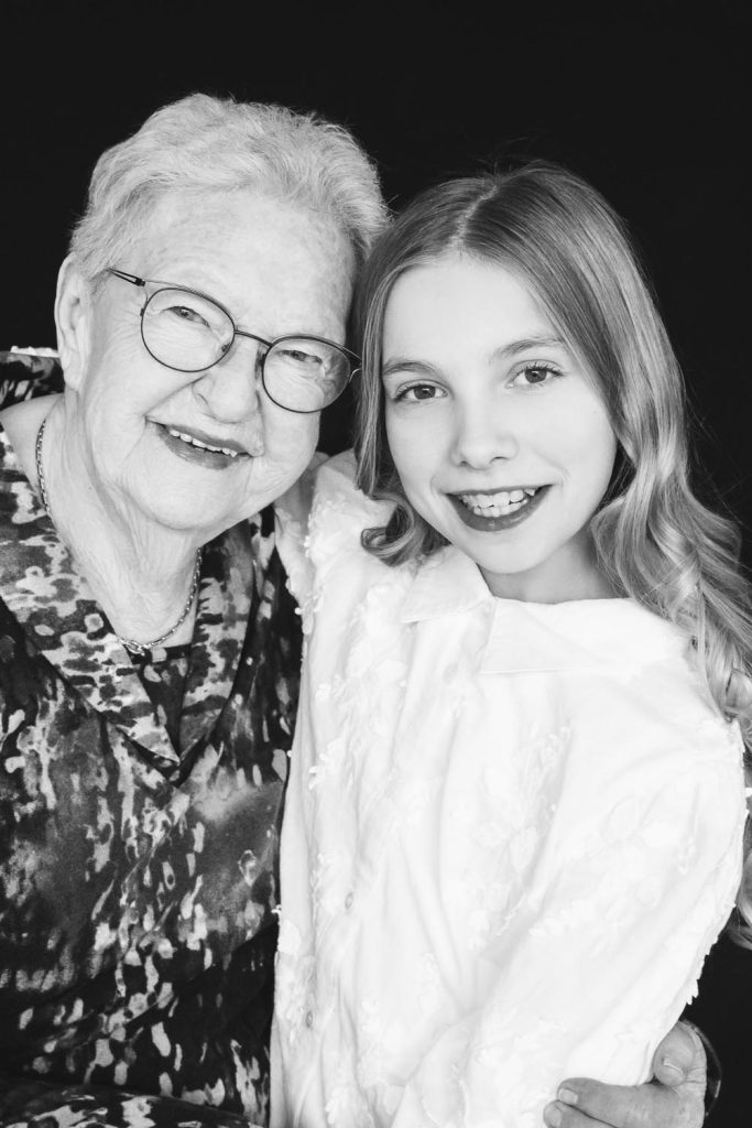 Black and white portrait of an elderly woman with glasses and a young girl smiling together with their arms around each other against a black background.