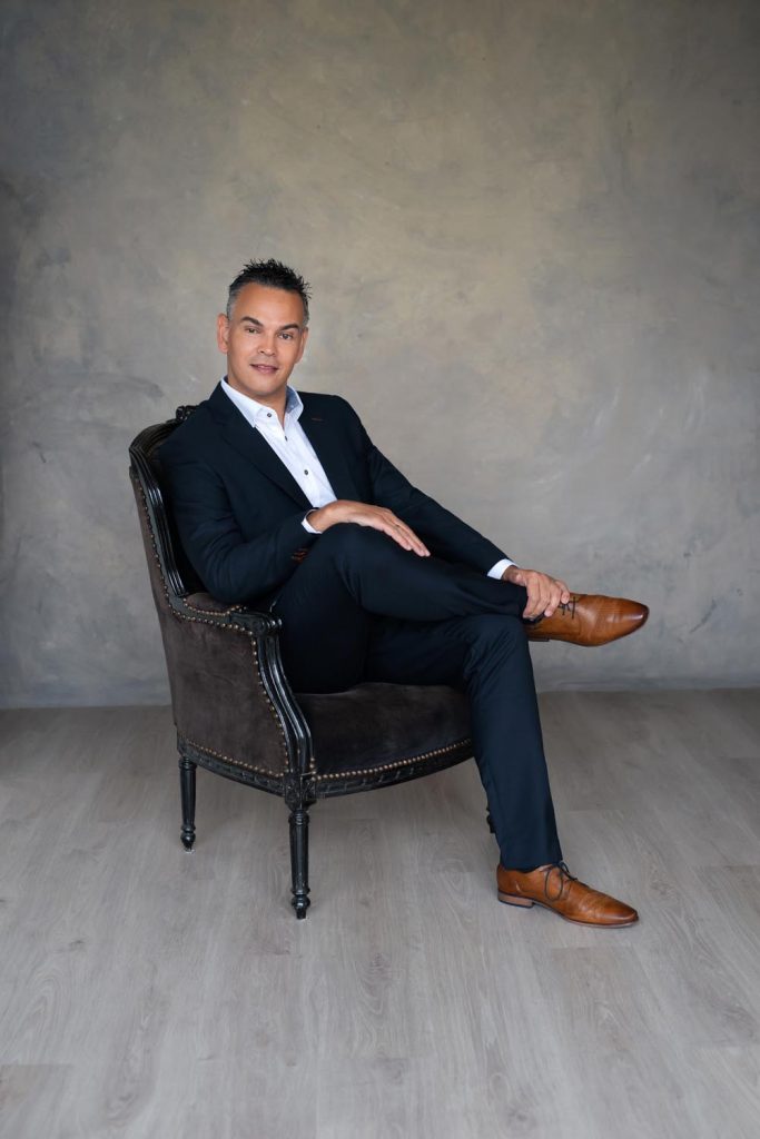 Man in a black suit with a white shirt sitting cross-legged on a dark vintage armchair against a plain textured wall and light wooden floor, wearing brown dress shoes and smiling slightly.