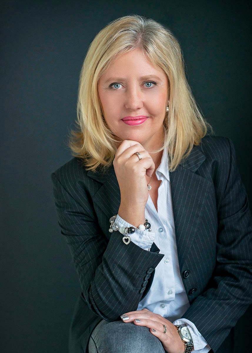 Blonde woman with blue eyes wearing a dark pinstripe blazer over a white shirt, posing thoughtfully with her hand near her chin against a dark background.