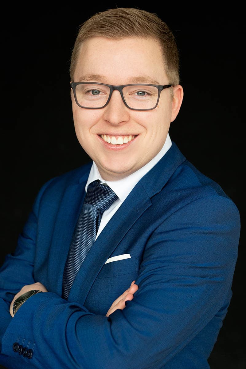 Smiling young man with short light brown hair and glasses wearing a blue suit, white shirt, and dark tie, posing with folded arms against a black background.