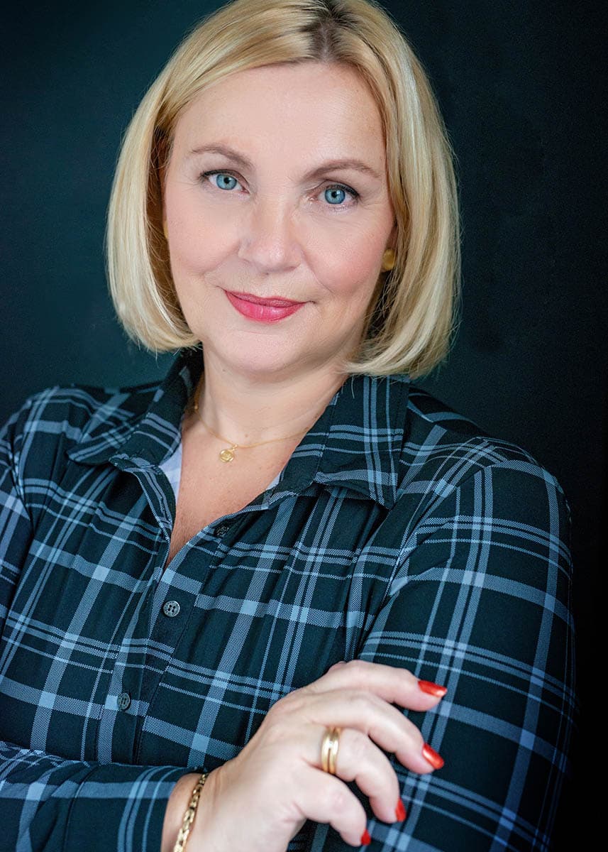 Close-up portrait of a middle-aged woman with blonde hair wearing a dark green and black plaid shirt, small gold necklace, and gold bracelet, smiling softly with arms crossed against a dark background.