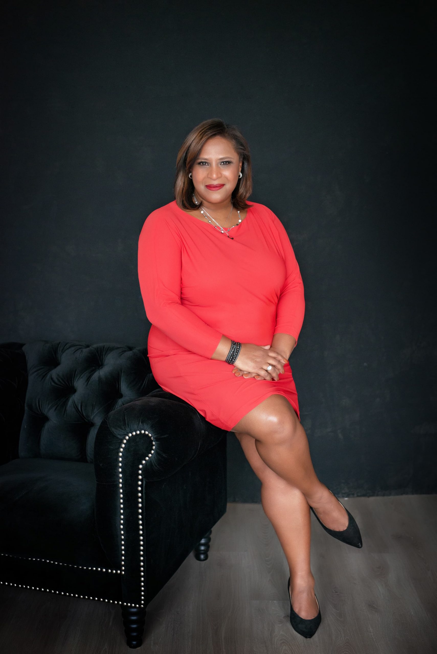 Woman sitting on the armrest of a black tufted sofa against a black wall, wearing a red dress and black heels, with short hair and a necklace, smiling at the camera.