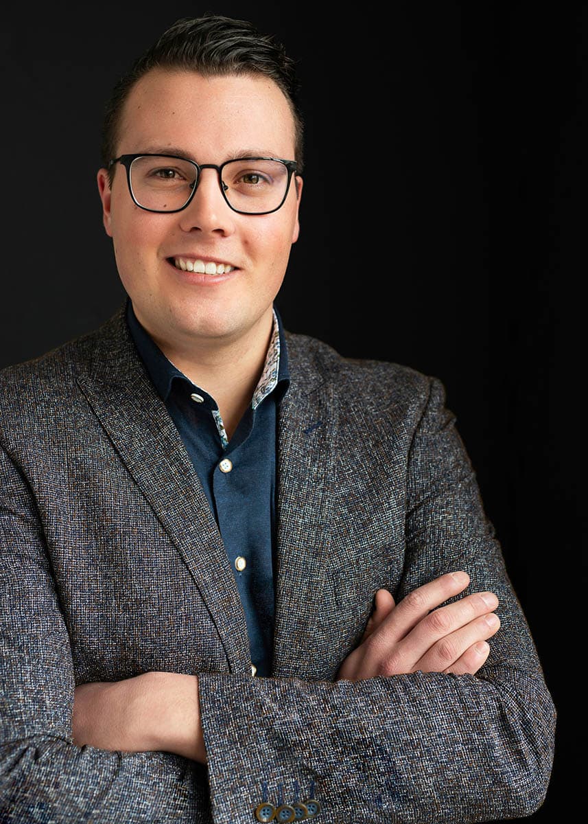 Smiling man with short dark hair and glasses wearing a dark blue shirt and textured gray blazer with arms crossed against a black background.