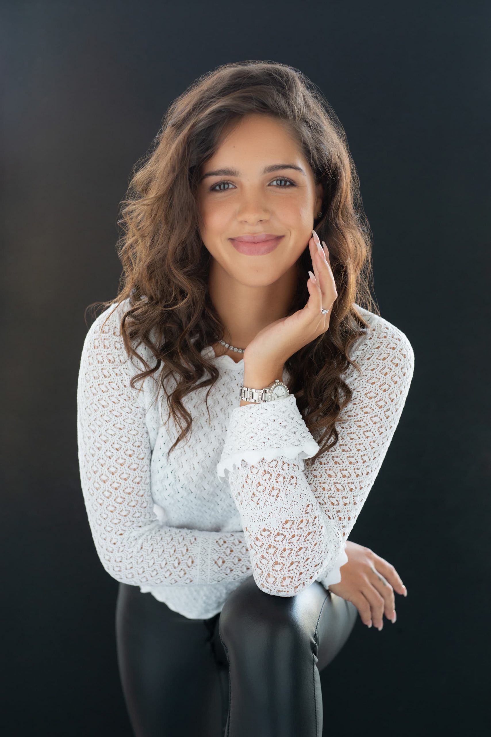 Young woman with curly brown hair, wearing a white lace long-sleeve blouse and black pants, smiling with her left hand resting on her cheek and her right hand on her knee against a dark background.
